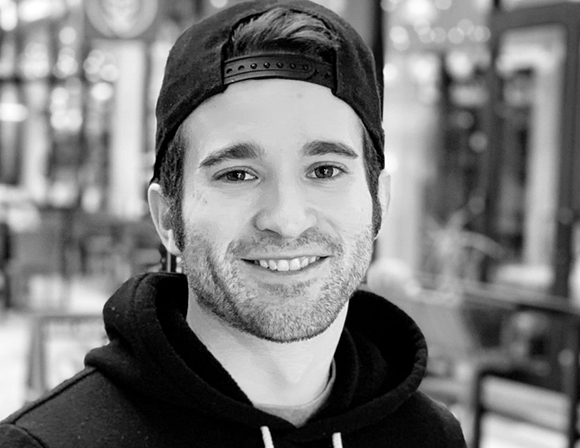 Black and white close-up portrait of a smiling man wearing a backwards baseball cap and a dark hoodie.
