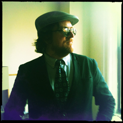 A man wearing a flat cap, glasses, and a suit stands near a bright window.