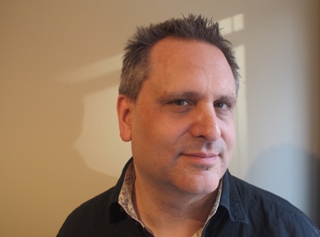 A man with short dark hair wearing a black button-down shirt against a neutral wall.