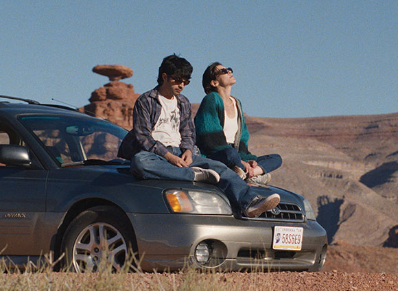 Two people sit on the hood of a dark SUV overlooking a desert canyon under a clear blue sky.