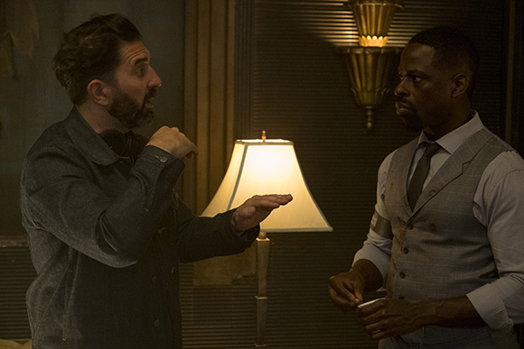 A man in a dark shirt gestures toward a man in a grey vest and tie in a dimly lit room.