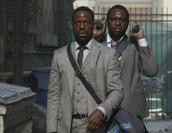 Two men in grey suits stand outdoors; one man in front looks forward while the man behind holds two handguns.