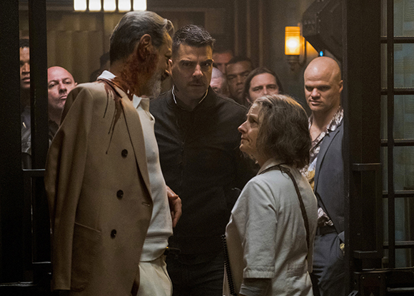A group of people stand in a dimly lit room, including a man in a tan coat with blood on his shoulder.