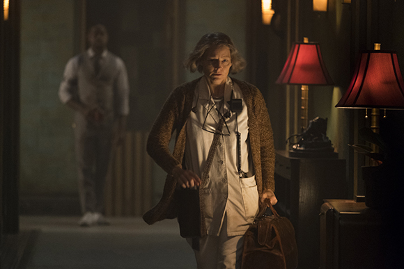 An older woman in a white uniform and brown cardigan walks through a dark hallway toward the camera.