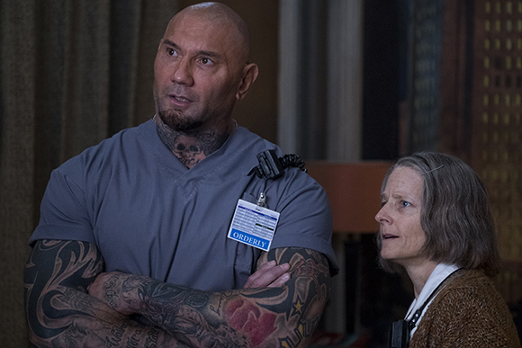 A bald, tattooed man in blue scrubs looks away while a woman in a brown sweater looks up at him.