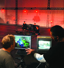 Film crew watching two monitors showing a scene with a blue figure in a red-lit studio.