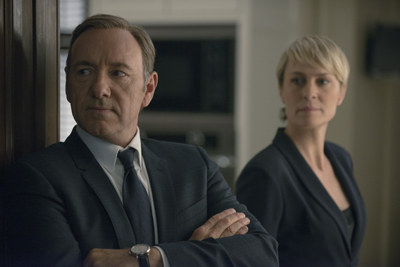 A man in a dark suit and tie with arms crossed stands in front of a woman in a dark blazer.