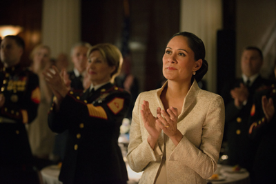 A woman in a cream blazer claps her hands during an indoor formal gathering.