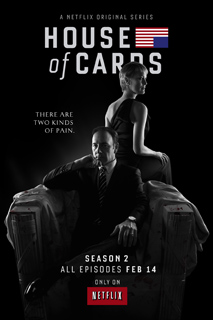 A man in a suit sits in a large chair while a woman stands behind him against a black background.
