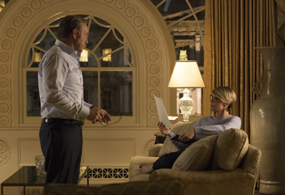 A man in a light shirt stands facing a woman sitting on a sofa holding papers in a dimly lit room.