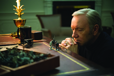 A man rests his chin on his hands while staring at small toy soldiers on a wooden desk.