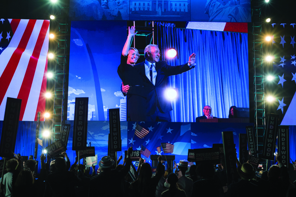Two people wave from a large stage in front of a blue backdrop and American flags.