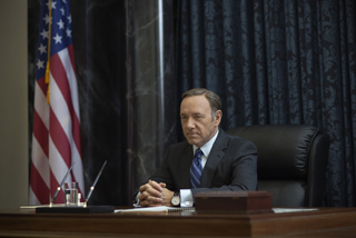 A man in a dark suit and blue tie sits at a wooden desk before an American flag and dark curtains.