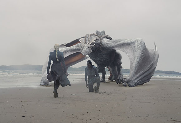 A woman in a dark coat stands on a sandy beach facing a massive grey dragon and a kneeling man.