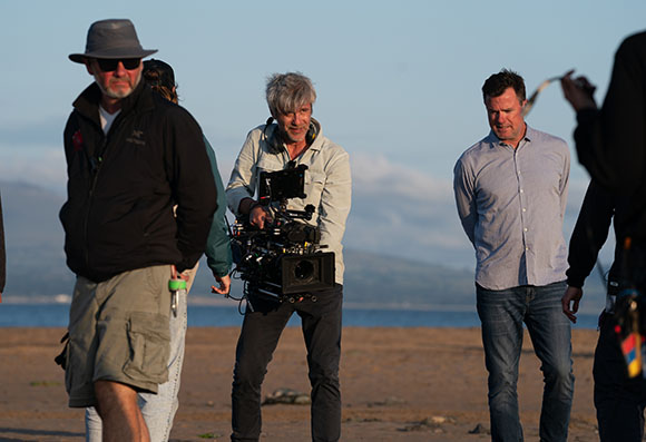 A film crew with a large camera and crew members walking on a sandy beach during twilight.