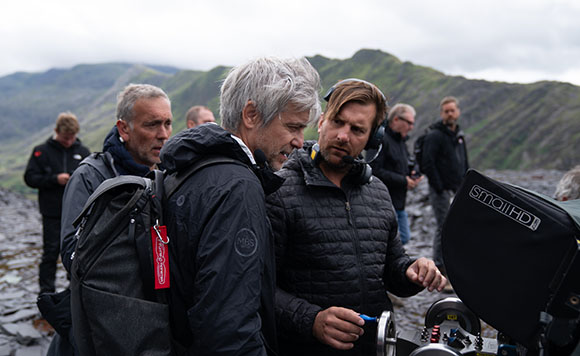 A group of men in outdoor gear discuss work near a professional camera in a rocky, mountainous area.