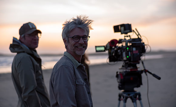 A man smiles at the camera on a beach next to a video camera and another man at sunset.