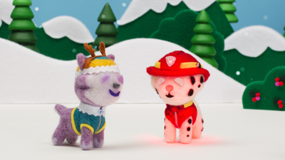 Two felt animal figures, one purple with antlers and one white with spots and a red hat, in a snowy scene.