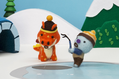 Two felt creatures, one orange and one blue, fishing on a white surface with a snowy landscape background.