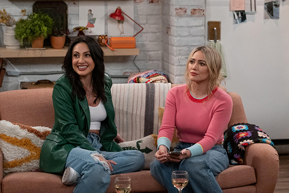 Two women sit on a brown couch; one wears a green leather jacket, the other a pink sweater.
