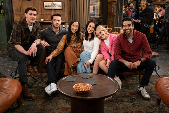 Six diverse friends smiling and sitting together on a couch and stools in a warm, dimly lit lounge.