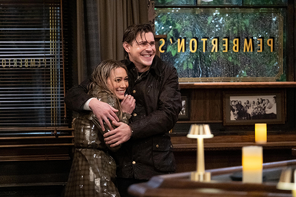 A man smiles while hugging a woman in a dark, wood-paneled room with a window in the background.