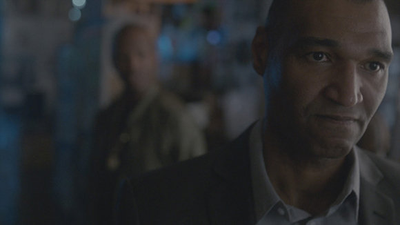 A man in a suit looks thoughtfully to the side in a dimly lit, blurred indoor setting.