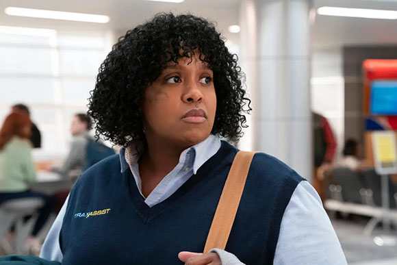 A woman with curly black hair wearing a blue collared shirt and a navy vest looks thoughtfully to the side.