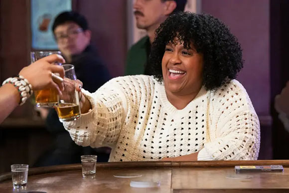 A smiling woman with curly black hair raises a glass to toast with others at a wooden bar.