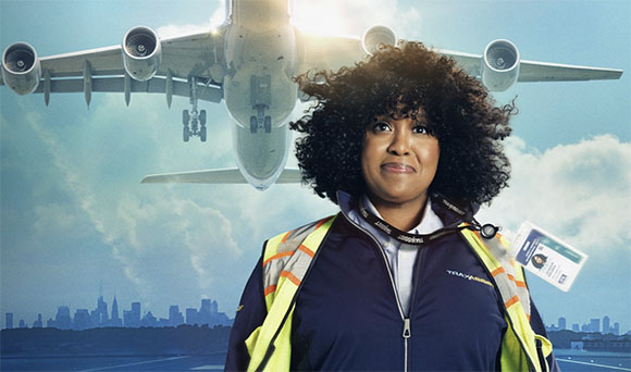 A woman with dark curly hair wearing a high-visibility vest and airport uniform, with a large plane flying overhead.