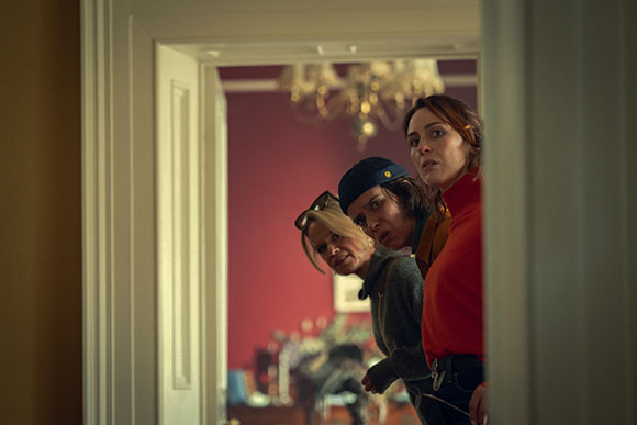 Three women look cautiously through an open doorway with concerned facial expressions.