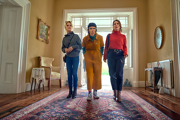 Three women stand in a room with wooden floors and a patterned rug, looking forward with serious expressions.