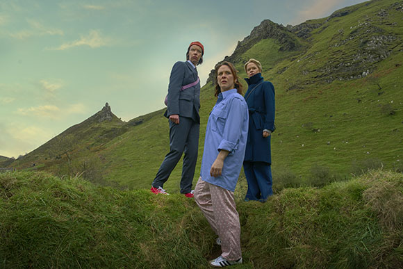 Three people in various colored clothing stand on a grassy slope with mountains under a cloudy sky.