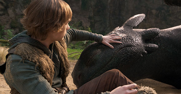 A young man with light brown hair sits on the ground, gently resting his hand on a large, dark-scaled dragon.
