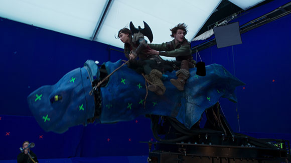 Two actors sit on a large blue mechanical dragon prop against a blue screen backdrop.