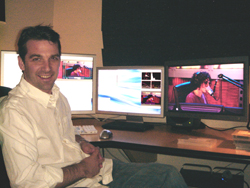 A smiling man in a white shirt sits at a desk with three computer monitors displaying various images.