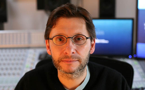 A man with short brown hair and round glasses looking at the camera in a studio.