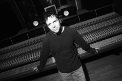 A man in a dark sweater leans against a large professional audio mixing console in a studio.