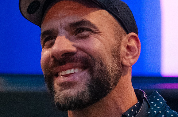 A smiling man with a dark beard and a black baseball cap looking off to the side.