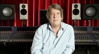 A man in a light blue striped shirt sits before a large mixing console and studio speakers against red curtains.