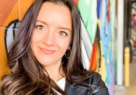 A woman with long dark hair and hoop earrings smiling in front of a vibrant, multicolored mural.