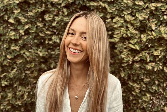A smiling woman with long blonde hair wearing a white shirt against a leafy green background.