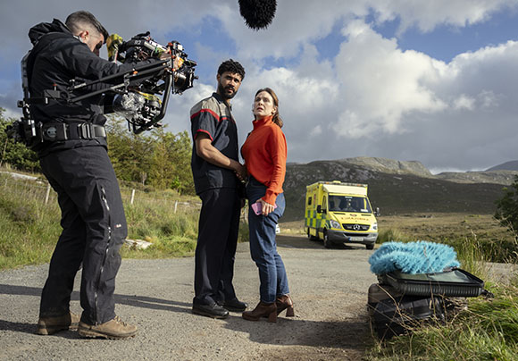 A camera operator films a man and woman standing on a road near an ambulance and mountains.