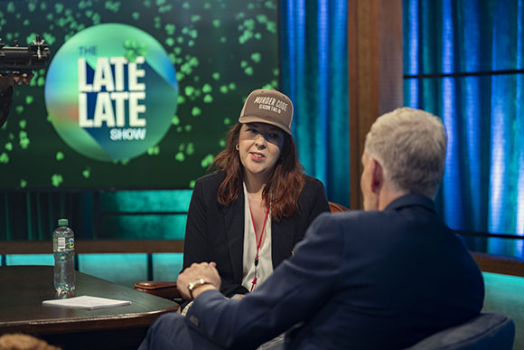 A woman in a dark blazer and baseball cap sits across from a man in a suit on a television set.