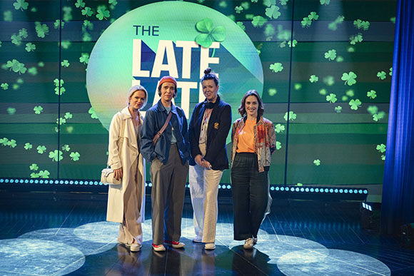 Four women posing together on a television set with a green clover-themed background.