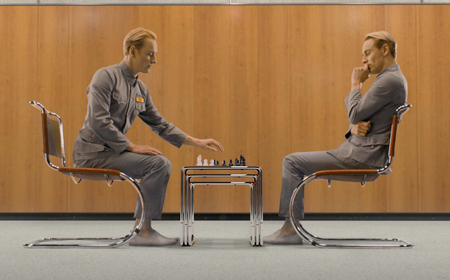 Two men in matching grey jumpsuits sit opposite each other playing chess at a small table.