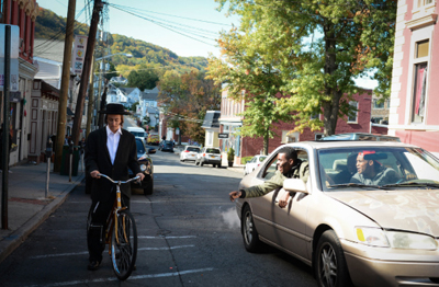 A man in a black suit and hat rides a bicycle past a tan car on a street lined with buildings and trees.
