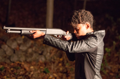 A person in a black jacket aims a shotgun toward the left in a dark, wooded setting.