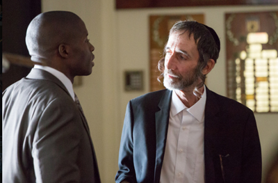 A Black man in a grey suit looks at a white man wearing a yarmulke and a black blazer.