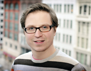 A man with short brown hair and black-rimmed glasses wearing a striped sweater, smiling against a blurred city backdrop.